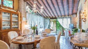 A dining area with a wooden table set for four, surrounded by white chairs. A chandelier hangs above, and a television is visible in the background. - Borgo Paradiso 4 2 in Ciciano (Chiusdino)