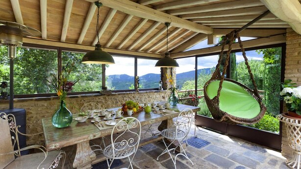 A rustic outdoor dining area with a wooden table, white metal chairs, hanging lamps, and a view of mountains through large windows. - Borgo Paradiso 4 2 in Ciciano (Chiusdino)