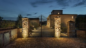 A stone-walled building with a metal gate, a paved driveway, and a fenced area with trees.