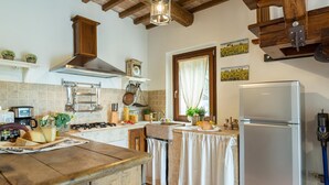 A kitchen with a stove, sink, and refrigerator. There are wooden cabinets and a window with curtains. - Casale Montegiovi 6 4 (Castel del Piano)