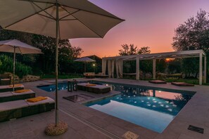Une piscine extérieure rectangulaire, dotée de chaises longues et de parasols, et entourée d’une pergola et d’arbres.