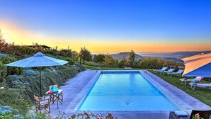 A rectangular pool with a clear blue water surface, surrounded by a paved deck. There are lounge chairs and umbrellas arranged around the pool, and a table with chairs nearby. The background features a natural landscape with trees and a view of distant hills. - Casale Le Conce 6 (Castel del Piano)
