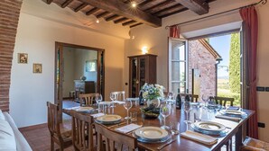 A dining area with a wooden table set for six, surrounded by chairs. There is a cabinet with glass doors and a window with a view of greenery outside. - Il Fitto 10 in Cortona (Cortona)