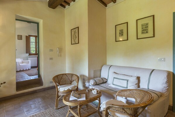 A cozy living room with a sofa, two wicker chairs, and a small table with a teapot and cups. There are framed pictures on the wall and a window with a view of another room.