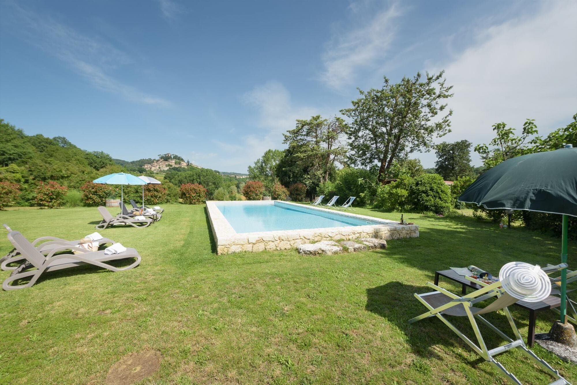 A rectangular pool with a stone border, surrounded by a grassy lawn. There are lounge chairs and umbrellas, and a view of a hill in the background.