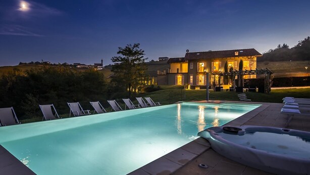 Une piscine avec des chaises longues, un jacuzzi et une maison dont l’extérieur est bien éclairé au crépuscule.