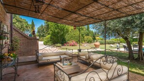 A covered outdoor seating area with cushioned benches and a table, surrounded by greenery and a view of the sea. - Casale Dei Girasoli 14 (Cortona)