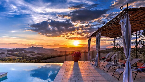 Piscina infinita con vista al atardecer, mobiliario exterior y una pérgola con cortinas.