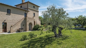 A stone building with a central tower, a covered outdoor seating area, and a well-maintained lawn. - Casale Cinque Valli 8 (Cortona)