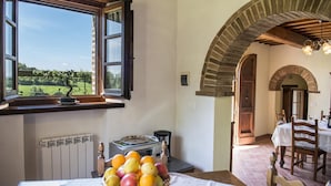 A room with a large window overlooking a garden, a dining area with a table set for two, and a radiator under the window. - Casale Cinque Valli 8 (Cortona)