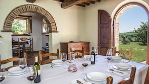 A dining area with a table set for four, surrounded by wooden chairs, a wooden cabinet, and a view of a vineyard through an arched doorway.