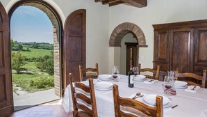 A dining room with a table set for four, large wooden chairs, and a view of a green landscape through an open archway. - Casale Cinque Valli 8 (Cortona)
