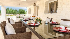 A dining area with wicker furniture, a glass table, and a view of trees and a garden. - White Stone House 6 1 (Ostuni)