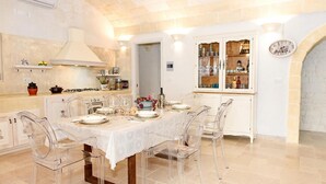 A dining area with a table set for four, clear chairs, a white tablecloth, and a kitchen area with a stove and cabinets. - White Stone House 6 1 (Ostuni)