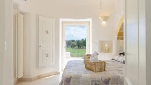 A bedroom with a large bed, a wooden chest of drawers, a bedside table with a lamp, and a view of a green field through an open door. - White Stone House 6 1 (Ostuni)