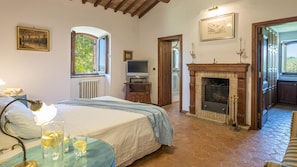 A bedroom with a bed, bedside table, lamp, and a view of a fireplace and kitchen.