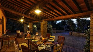 An outdoor dining area with stone pillars, wooden furniture, and a view of the garden at dusk. - Virginia 8 in Gavorrano (Scarlino)