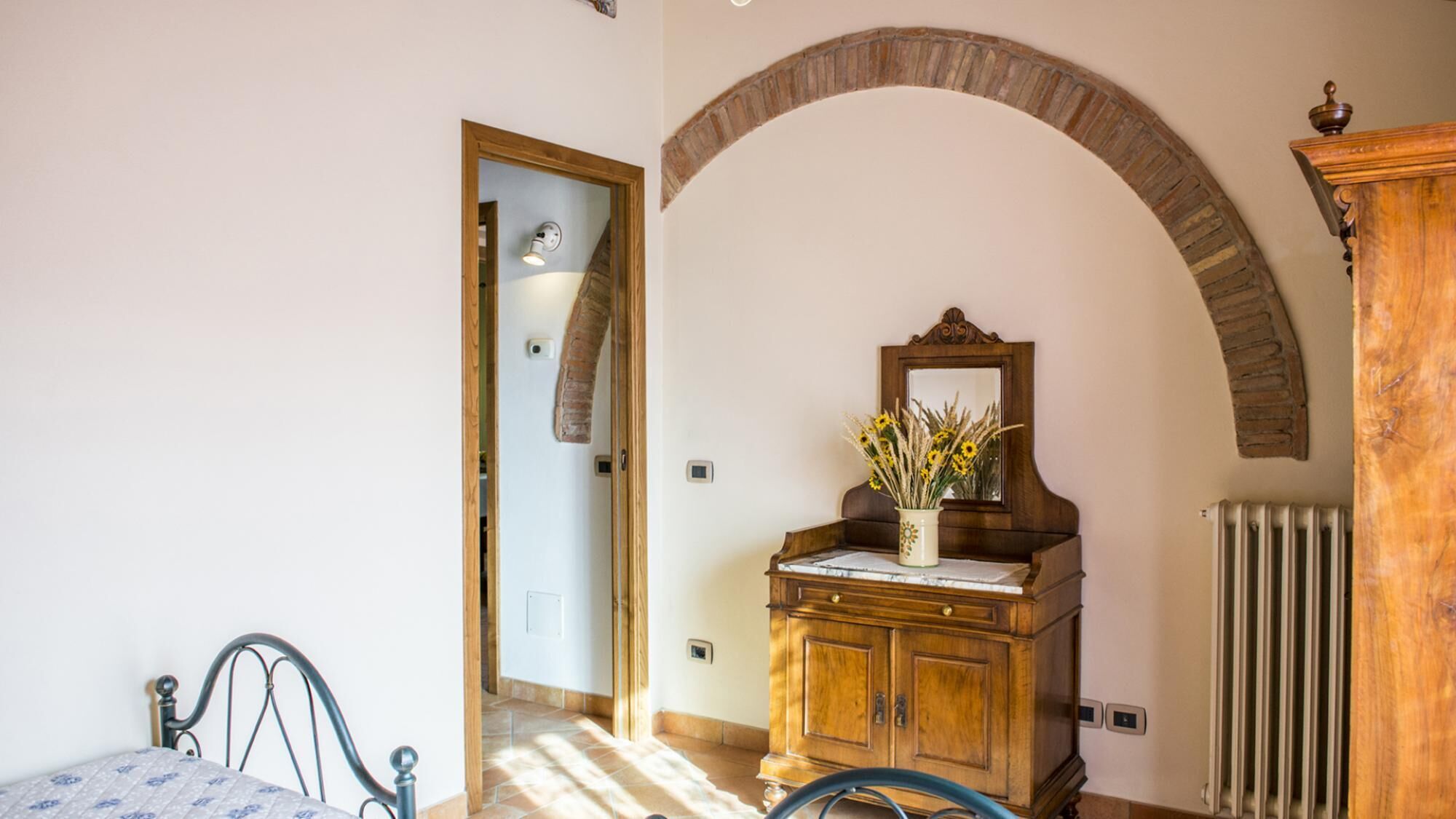 A bedroom with a bed, a wooden dresser with a mirror, a radiator, and an arched doorway.