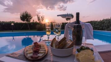 Un tavolo con bottiglia di vino, bicchieri, pane, formaggio e olive, affiancato a una piscina al tramonto.