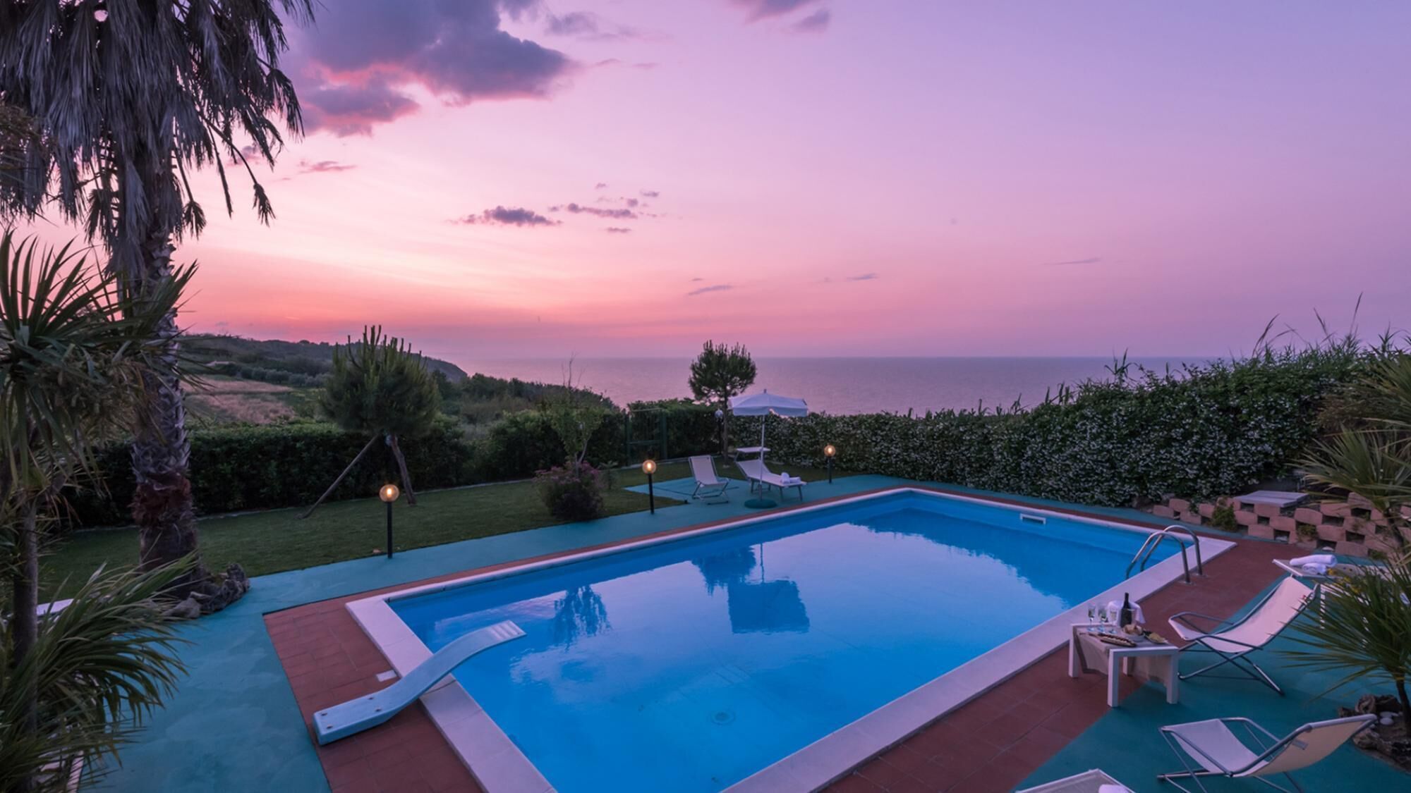 Una piscina con vista sul mare al tramonto, immersa nel verde e circondata da sdraio.
