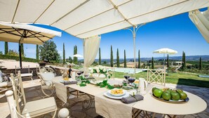 Un espace de restauration en plein air avec une table dressée pour un repas, entourée de chaises et de parasols, offrant une vue sur une piscine et le paysage environnant.