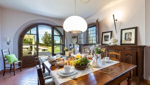 Un coin repas avec une table en bois dressée pour le petit-déjeuner, de grandes fenêtres donnant sur l’extérieur et un lustre au-dessus.