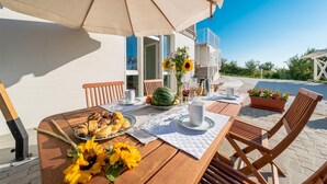 A wooden table with a white tablecloth, surrounded by wooden chairs, under a beige umbrella. There are sunflowers, a watermelon, and a plate of pastries on the table.