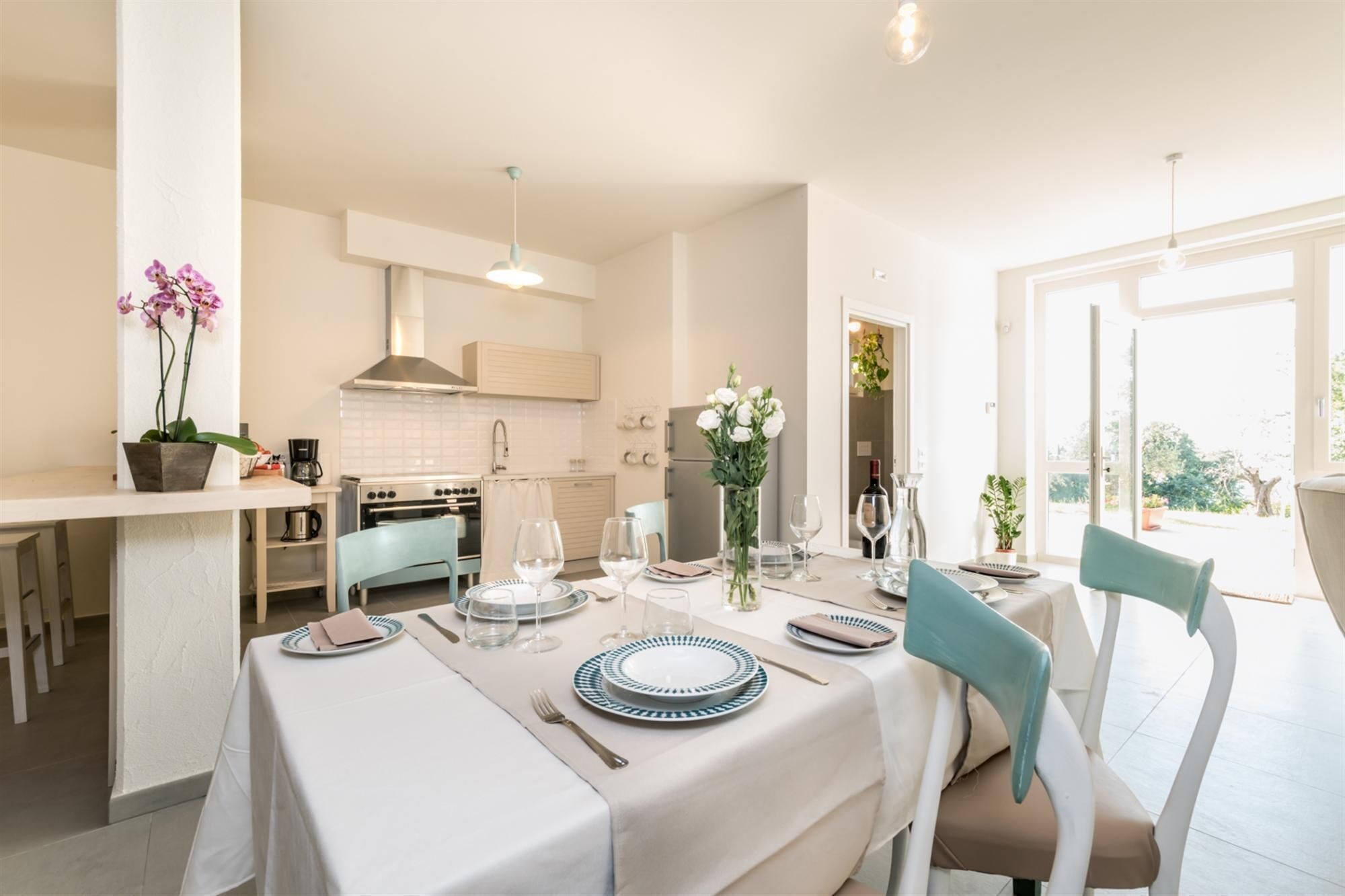 A modern kitchen with white cabinets, a stove, and a sink. There is a dining area with a table set for two, featuring plates, glasses, and a vase of flowers.