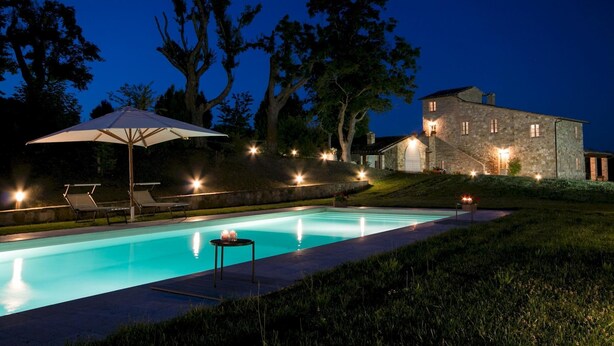 A nighttime view of a swimming pool with lounge chairs and an umbrella, surrounded by stone buildings and trees. - Podere Pisinano 8 2 (Montepulciano)
