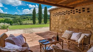A covered outdoor seating area with wicker furniture, a table with wine and glasses, and a stone wall. - Podere Pisinano 8 2 (Montepulciano)