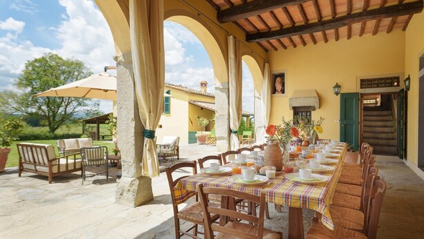 Un espace de restauration en plein air avec une longue table dressée pour le petit-déjeuner, entouré de piliers en pierre et surmonté d’une pergola en bois.