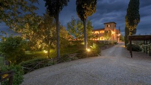 Un chemin de gravier mène à un bâtiment éclairé d’une lumière chaleureuse, entouré d’arbres et délimité par une clôture en bois.