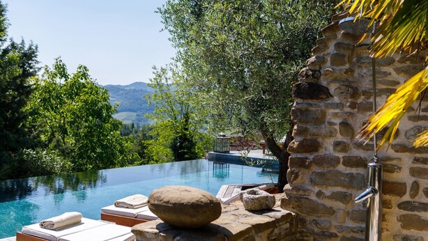 A pool with a stone wall and lounge chairs, surrounded by greenery and a view of hills. - Il Castellaccio 8 in Brisighella (Brisighella)