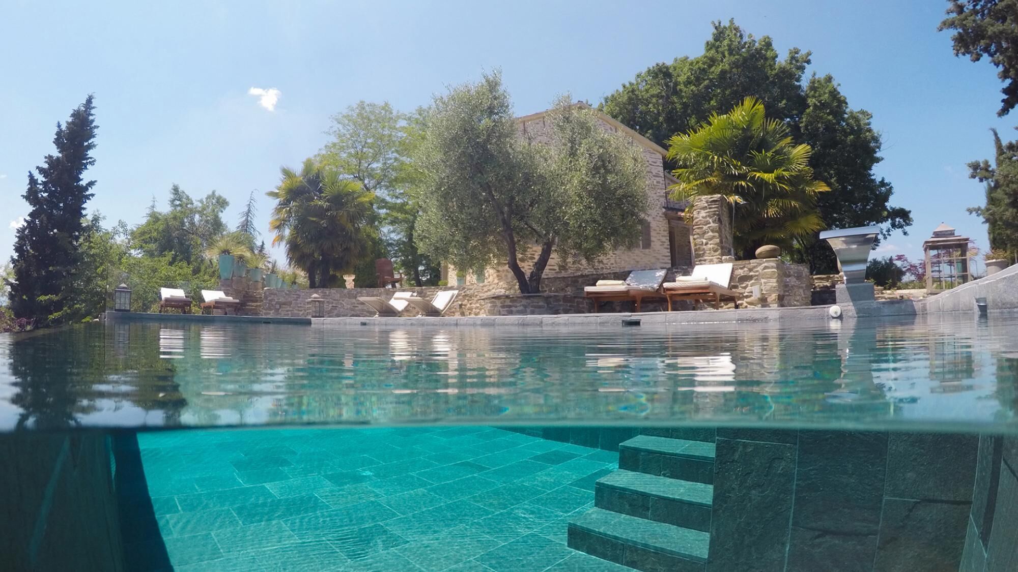 An infinity pool with clear blue water, surrounded by stone walls and trees. There are lounge chairs along the poolside.
