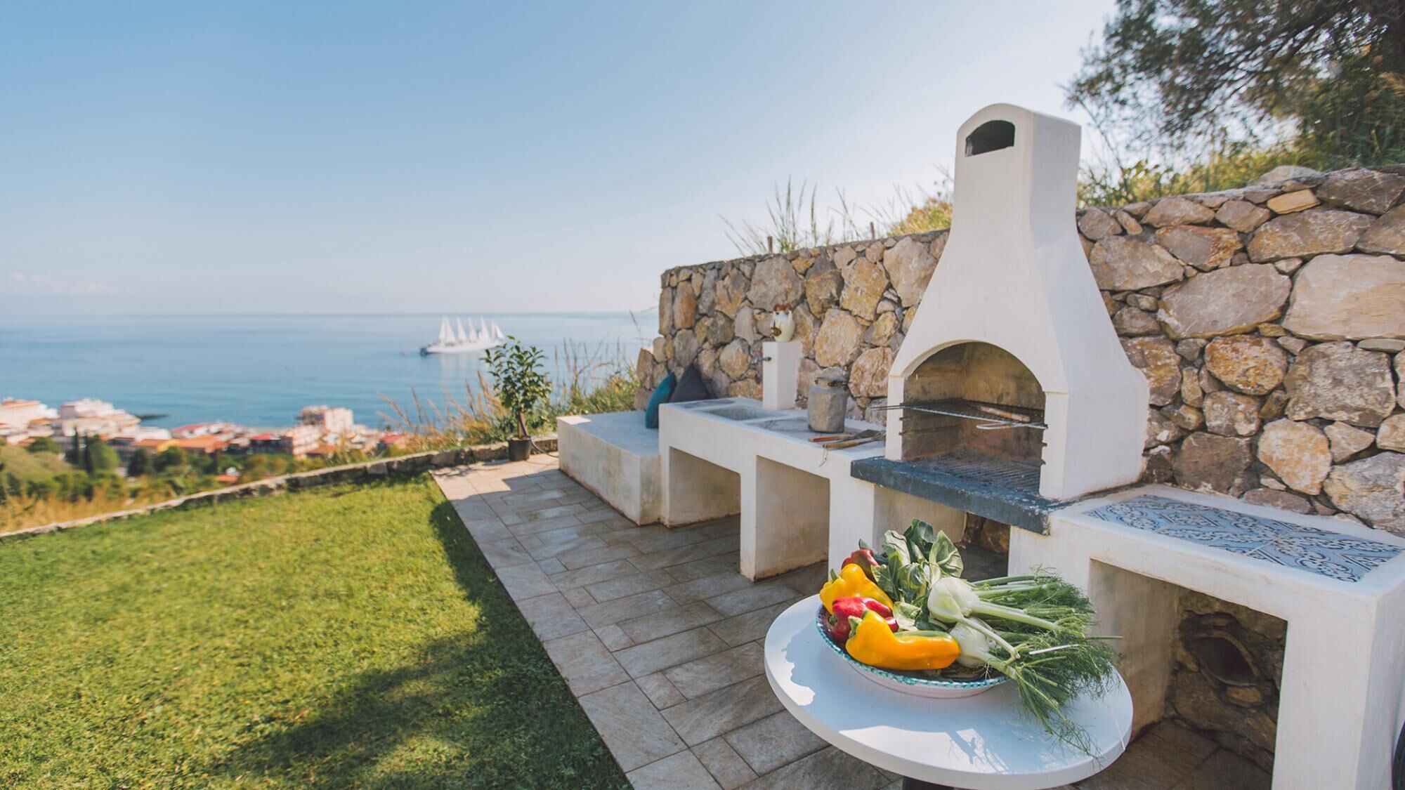 Une cuisine extérieure avec un mur en pierre en arrière-plan, une vue sur la mer et une table garnie de légumes frais.