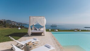 Un área de piscina exterior lujosa con una cama y sillones de descanso, vista despejada al mar y un velero a lo lejos.
