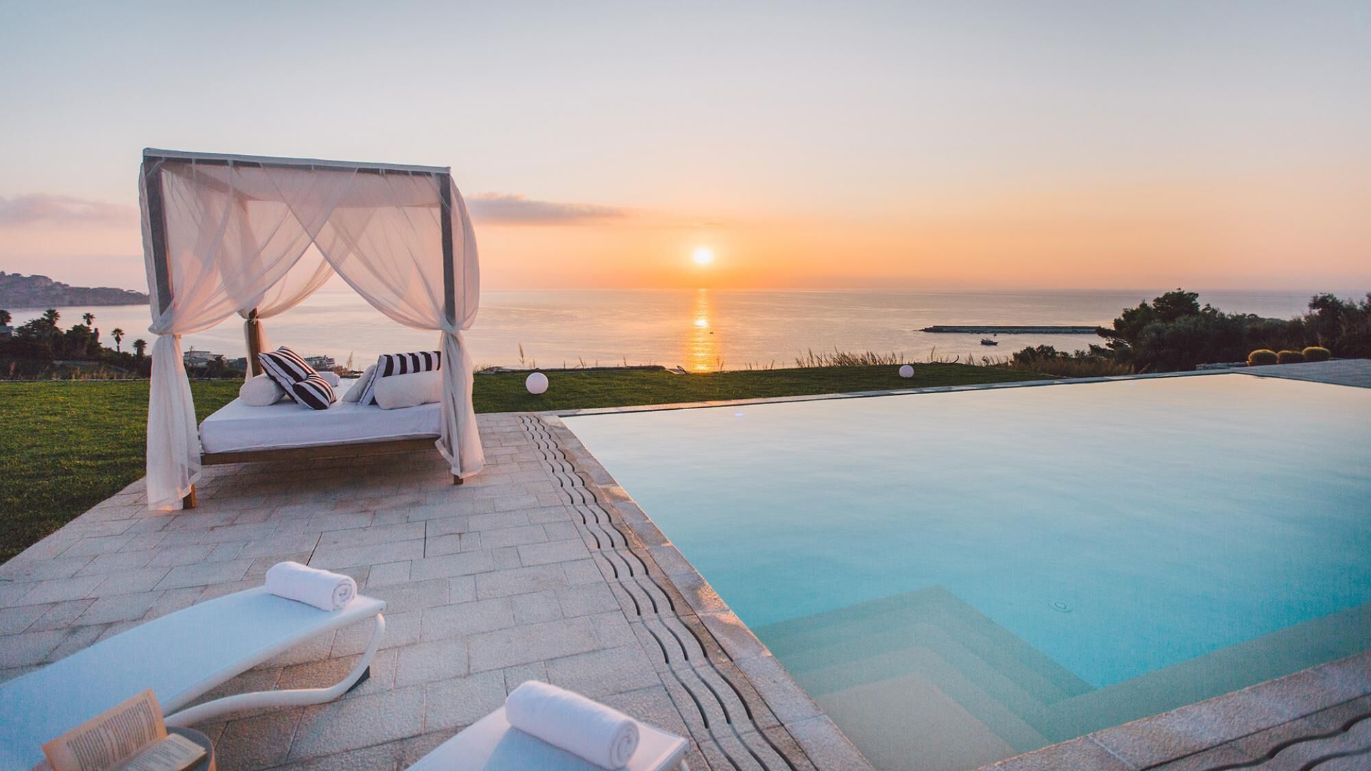 Un área junto a la piscina con una cama con dosel, sillones de descanso y vistas a la puesta de sol sobre el mar.