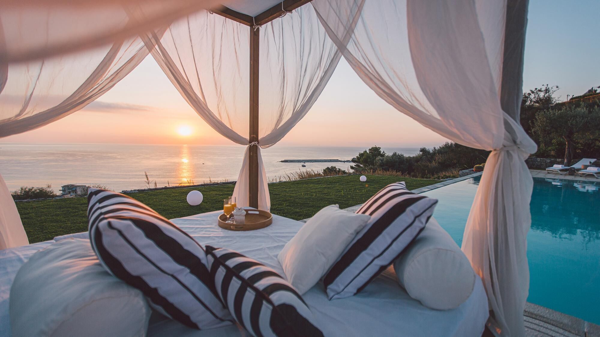 Ein Bett mit weiß-schwarz gestreiften Kissen, ein Tablett mit einem Glas Orangensaft und Blick auf den Sonnenuntergang über dem Meer.