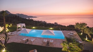 Zona de piscina con sillones y sombrillas, con vistas a un paisaje costero al atardecer.