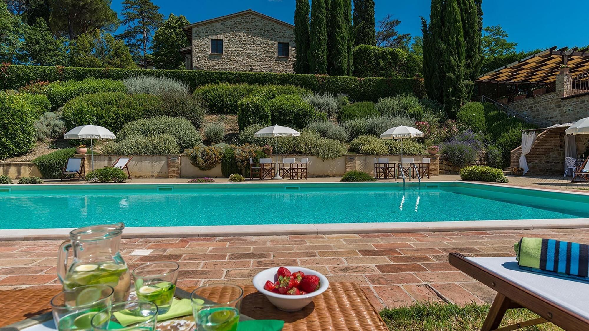 A poolside area with a stone house in the background, surrounded by lush greenery and tall trees.