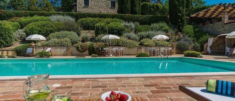 A poolside area with a stone house in the background, surrounded by lush greenery and tall trees.