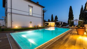 A swimming pool with a wooden deck, a white building with a large window, and a few trees in the background. - Villa Sole Al Molino A Vento 8 (Rosignano Marittimo)