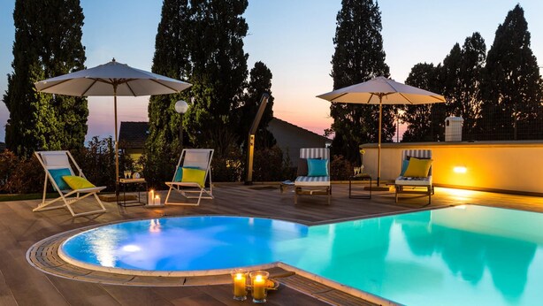 Un espace piscine agrémenté de chaises longues et de parasols, entouré de grands arbres et avec un coucher de soleil en toile de fond.