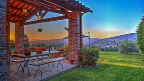 A covered outdoor seating area with a table and benches, surrounded by potted plants and a view of a town and mountains in the background. - San Rocco 4 in Montecchio (Montecchio)
