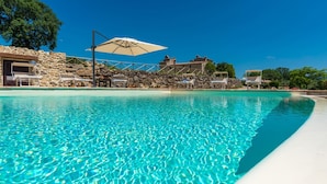 A clear blue pool with lounge chairs and umbrellas, surrounded by stone walls and buildings. - Santa Illuminata 20 12 (Massa Martana)