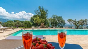 A poolside view with lounge chairs, a table with strawberries and drinks, and a scenic mountain backdrop.