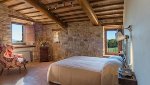 A rustic bedroom with a stone wall, wooden beams, a bed, a chair, and a window with a view. - Santa Illuminata 20 12 (Massa Martana)