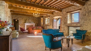 A rustic interior with exposed wooden beams, stone walls, and a fireplace. There are two upholstered armchairs, a sofa, and a dining table. - Santa Illuminata 20 12 (Massa Martana)