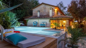A poolside area with a wooden deck, lounge chairs, and a stone building with a tiled roof.