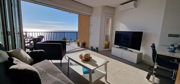 A modern living room with a sofa, a coffee table, a TV, and a balcony with a view of the sea.